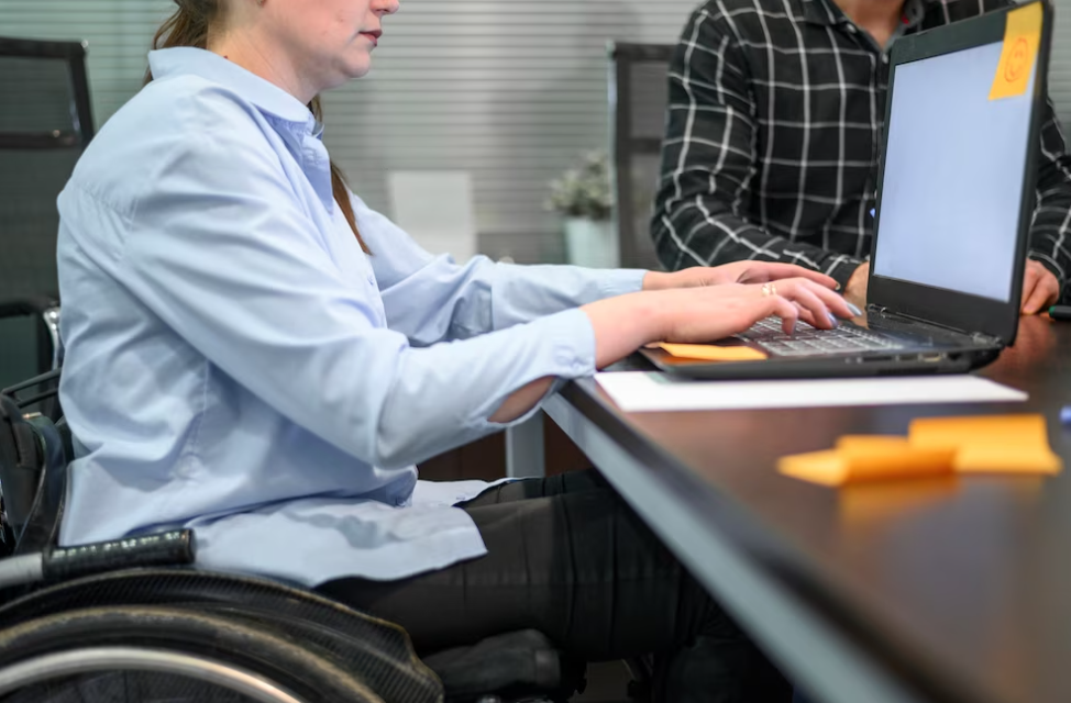 woman in the blue shirt in a wheelchair types on her laptop, man sits near her